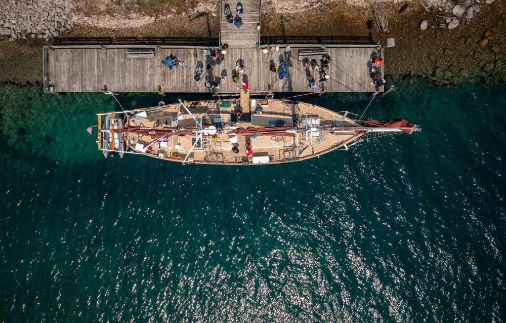 Aerial view of a sailboat docked at a pier with people on it, set in clear turquoise water.